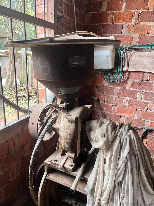 An old machine with a hopper beside a brick wall and window.