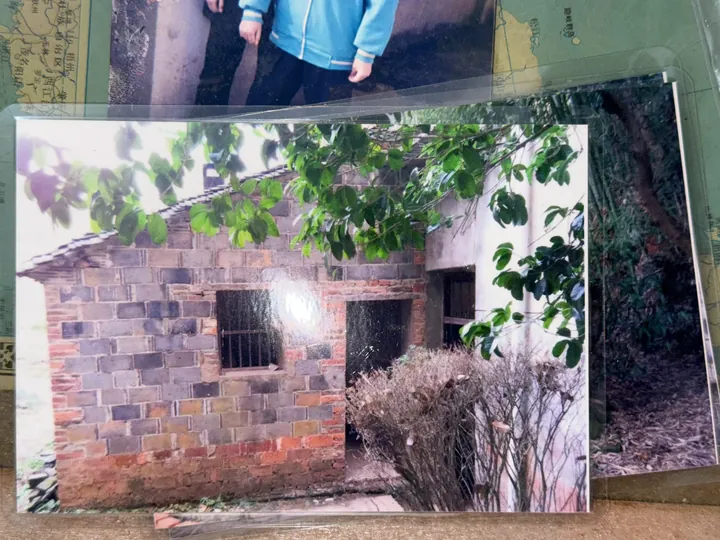 A photograph of an older printed image of a brick house, with branches and weeds in front.