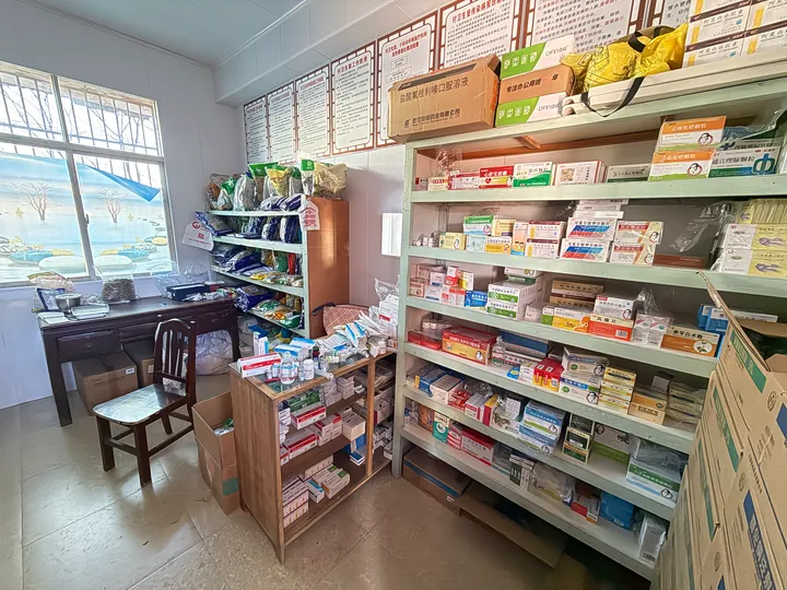 A room corner with medicine shelves, cardboard boxes, and a desk by the window.