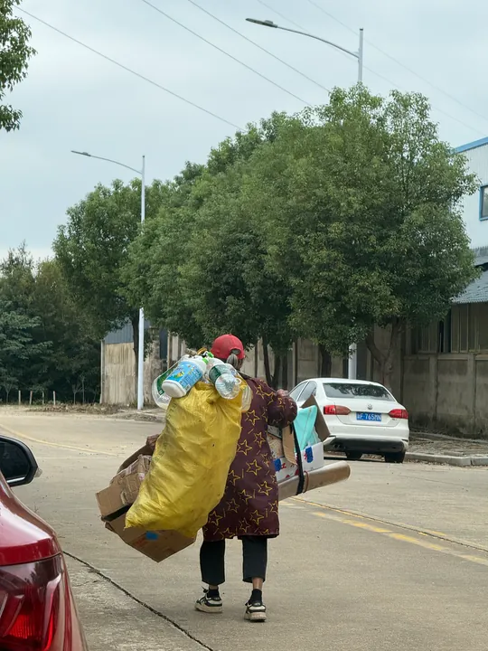 A person walking along a road carrying a large bag, bottles, and cardboard.