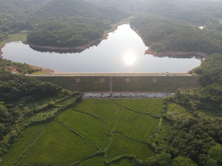 An aerial view of a reservoir, its dam, and the surrounding woods and fields.